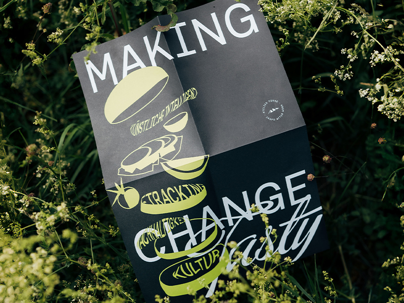 Künstlerisch gestaltetes Poster mit typografischer Collage und Illustrationen rund um Essen, Tracking und Kultur, designt von Atelier Türke, platziert in natürlicher Wiesenlandschaft. Ein kreatives Beispiel für experimentelles Plakat- und Brandingdesign.