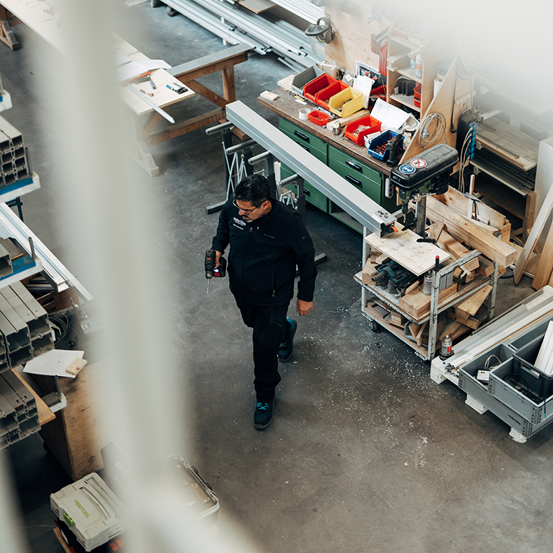 Mitarbeiter geht durch die Werkstatt von Atelier Türke mit Werkbänken, Maschinen und Materialien. Er hält eine Bohrmaschine in der Hand. Ansicht von schräg oben.