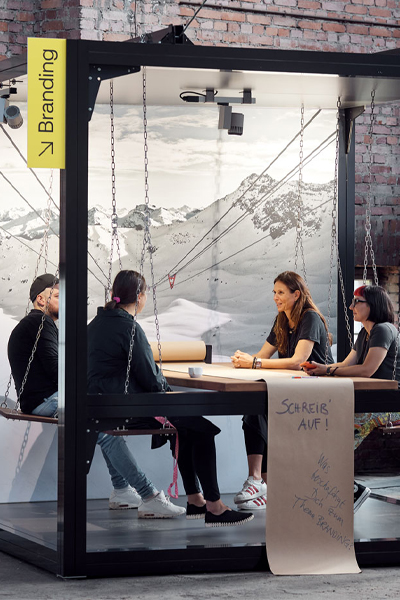 Pop-up meeting room in the industrial setting of the creative space in Atelier Türke’s Schwelhalle with a metal frame, a central table and people sitting on swings exchanging ideas, flanked by branding elements and an alpine image motif. This creates a temporary space for dialog, collaboration and brand communication.