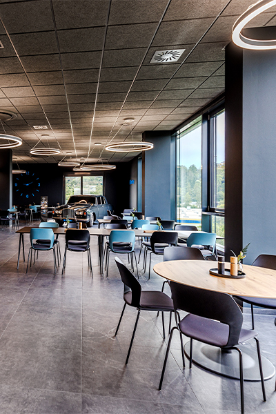 Großzügig gestalteter Arbeitsraum und Kantine mit runden Tischen, dunklen Stühlen, hohen Fenstern und markanten Ring-Leuchten. Atelier Türke schafft mit diese Einrichtung eine moderne Arbeitswelt mit viel Tageslicht, klarer Struktur und Raum für Austausch und Begegnung.