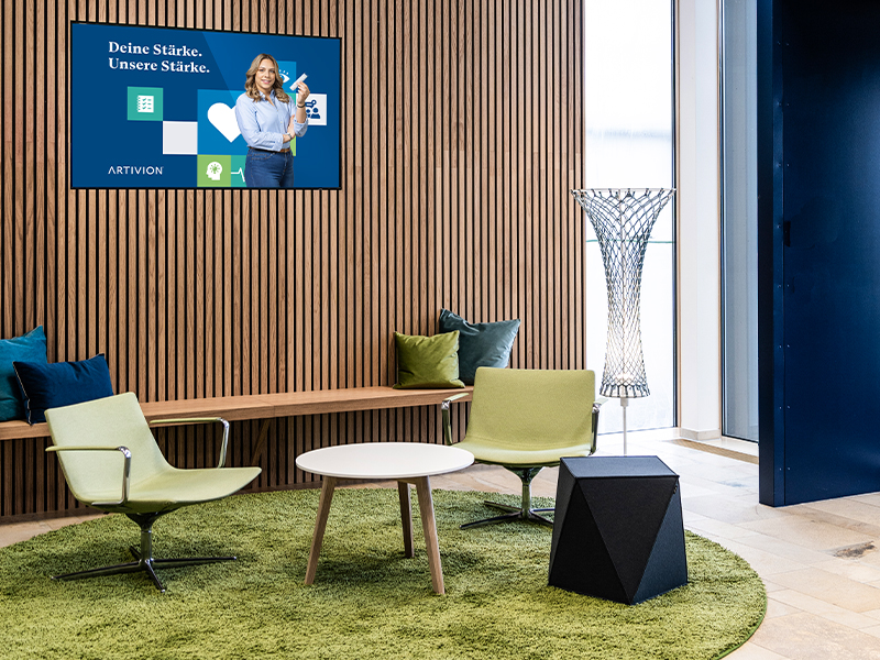 Einladende Sitzlounge mit grüner Teppichfläche, modernen Sesseln, rundem Beistelltisch und Holzlamellenwand mit integriertem Screen. Ein von Atelier Türke entworfener, ruhig gestalteter Aufenthaltsbereich für Austausch, Information und kurze Pausen.