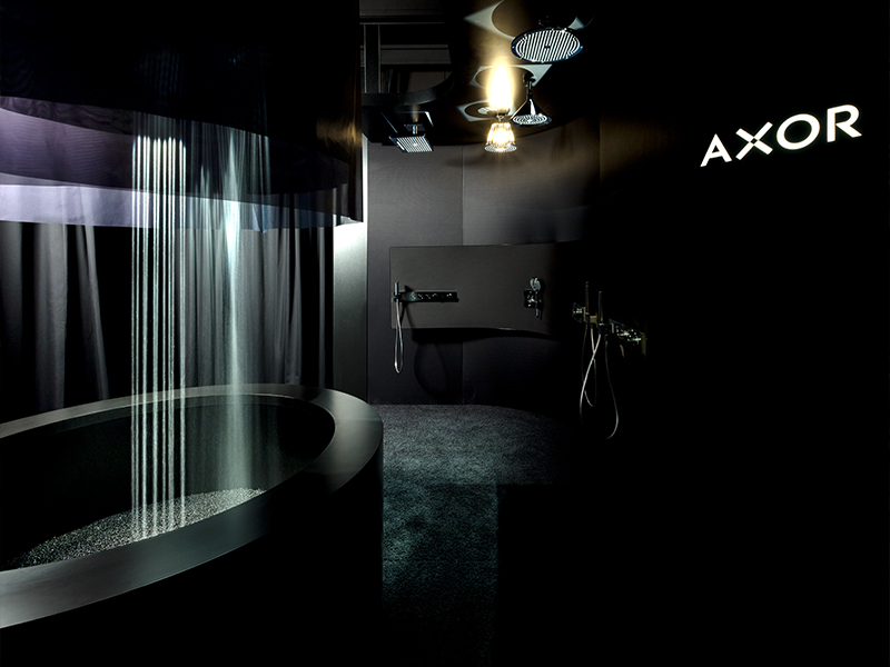 Dunkel inszenierter Axor-Showroom mit rundem Wasserbecken und sanft fallendem „PowderRain“-Wasserstrahl, akzentuiert durch gezielte Lichtinszenierung. Eine reduzierte Markenwelt, die das Element Wasser sinnlich erlebbar macht, und von Atelier Türke designt wurde.