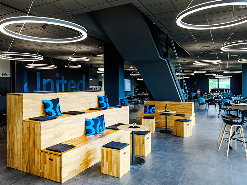 Modern gestalteter Arbeits- und Aufenthaltsbereich mit modularen Holz-Sitzstufen, Tischen und Stühlen, markanten Ring-Leuchten sowie offener Raumstruktur. Eine kommunikative Arbeitswelt für Austausch, Meetings und Pausen, von Atelier Türke entworfen und umgesetzt.