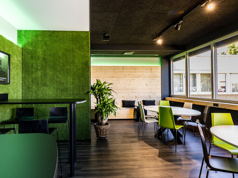 Modern gestalteter Aufenthalts- und Pausenbereich mit grüner Akzentwand in Moosoptik beim Unternehmen AKE mit hellen Holzelementen, runden Tischen mit grünen Stühlen und einem höherem Tisch mit schwarzen Barhockern. Das naturnahe Raumdesign von Atelier Türke lädt mit der entspannter Atmosphäre zum Austausch und zur Erholung ein.