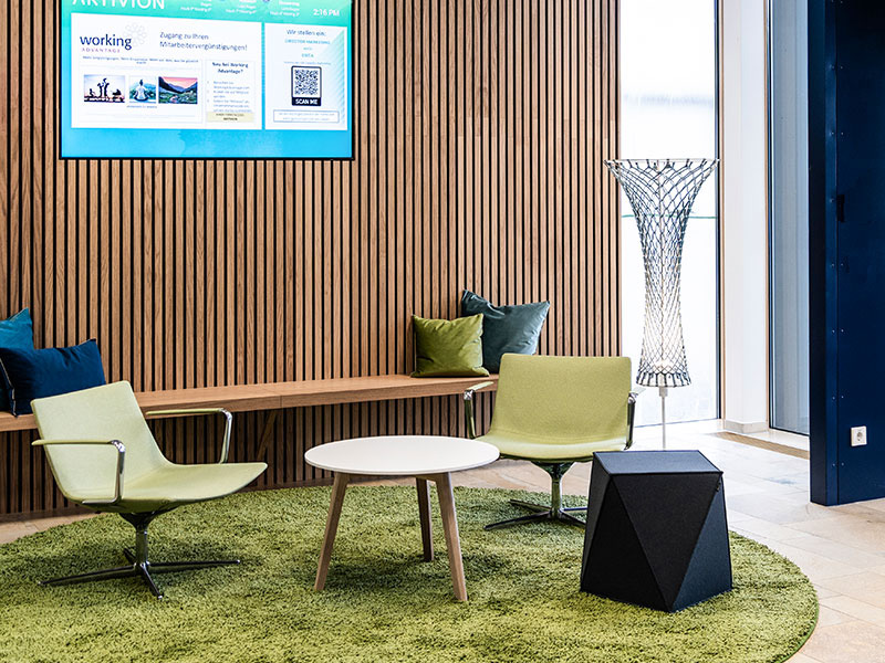 Modern gestaltete Sitzlounge als Arbeitswelt für Artivion mit grüner Teppelfläche, grünen Sesseln, einem runden Holztisch, Holzpaneelwand und integriertem Screen zur digitalen Markenkommunikation. Atelier Türke entwarf diesen einladenden Raum für Dialog und Aufenthalt im Unternehmen.