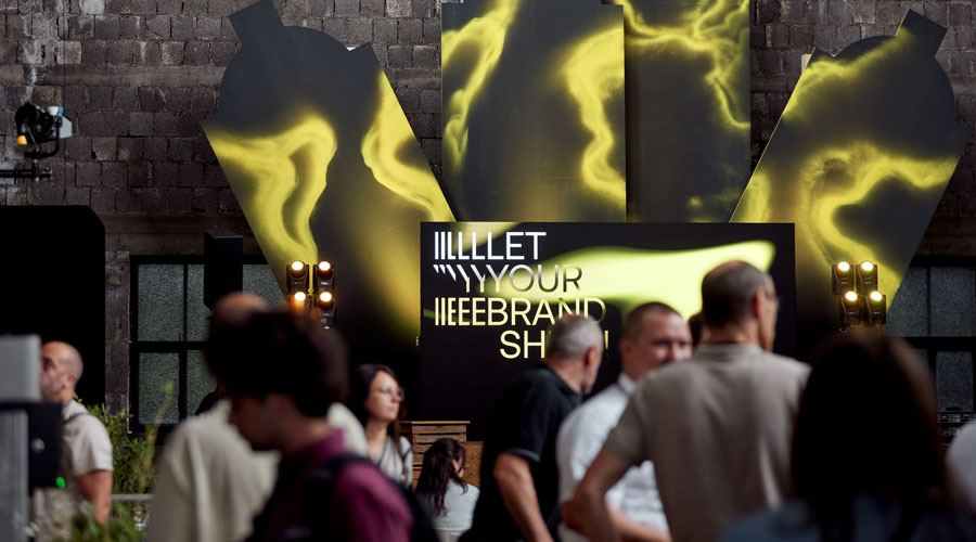 Inszenierter Messebereich bei der Hausmesse von Atelier Türke mit großformatigen, grafischen Elemente in kontrastreicher Farbwelt, leuchtender Markenbotschaft und Menschen im Vordergrund. Eine atmosphärische Markeninszenierung im Ausstellungsdesign mit starker visueller Wirkung.