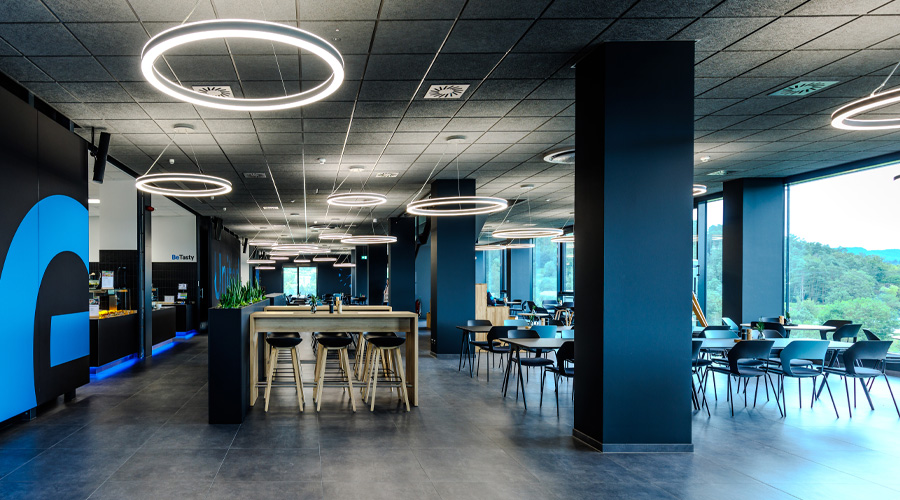 Großzügige, modern gestaltete Kantine als Raum zur Mehrfachnutzung mit offenen Arbeits- und Sitzflächen, hoher Decke, markanten Ring-Leuchten und klarer Markenfarbgebung von Bentley. Atelier Türke entwickelte diesen Raum für Austausch, Begegnung und gemeinsames Arbeiten.