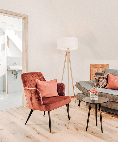 Gemütlich eingerichteter Innenraum des Hotels Krone Lautlingen mit einem Sessel, einem Sofa, einem Beistelltisch und einer Stehlampe vor dem hell eingerichteten Badezimmer. Warmes, wohnliches Raumdesign und Einrichtungskonzept von Atelier Türke.