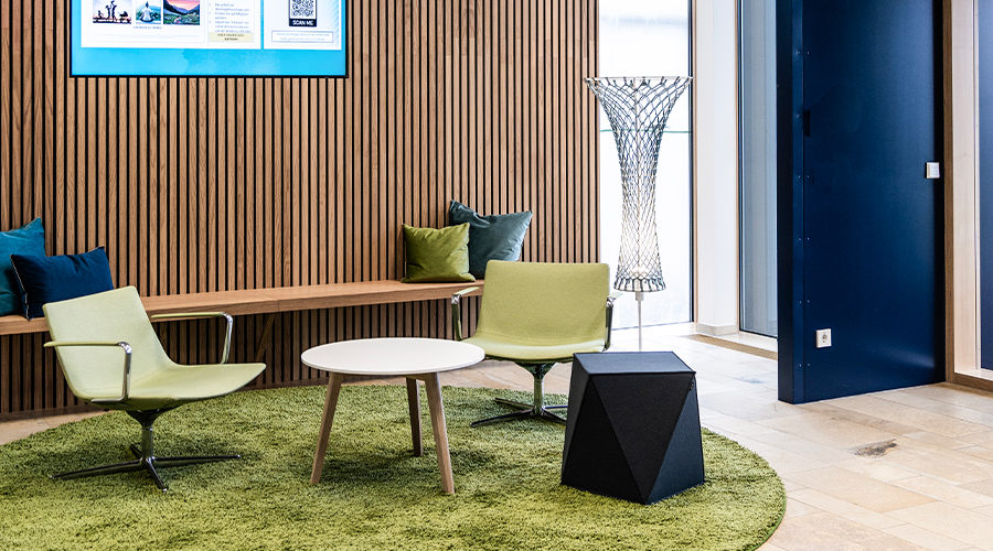 Modern gestaltete Sitzlounge als Arbeitswelt für Artivion mit grünem Teppich, grünen Sesseln, Holzpaneelwand und integriertem Bildschirm zur digitalen Unternehmenskommunikation. Atelier Türke entwarf diesen einladenden Raum für gemeinsames Arbeiten und Aufenthalt im Unternehmen.