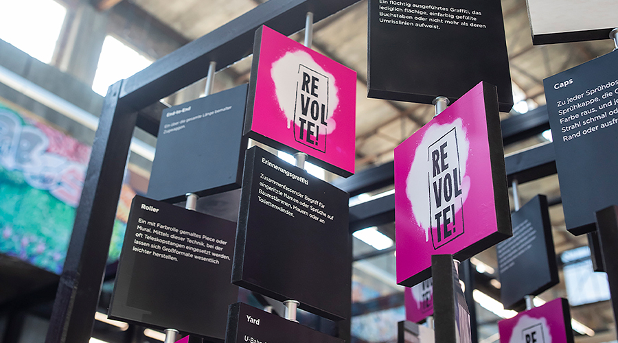 Hängende, modulare Präsentation mit pink-schwarzen Karten und der Aufschrift „REVOLTE!“ in der industriellen Umgebung des Kreativraums in der Schwelhalle von Atelier Türke. Das plakative, aufmerksamkeitsstarke Ausstellungsdesign zur Marken- und Themeninszenierung mit informativen Texttafeln ist interaktiv aufgebaut.